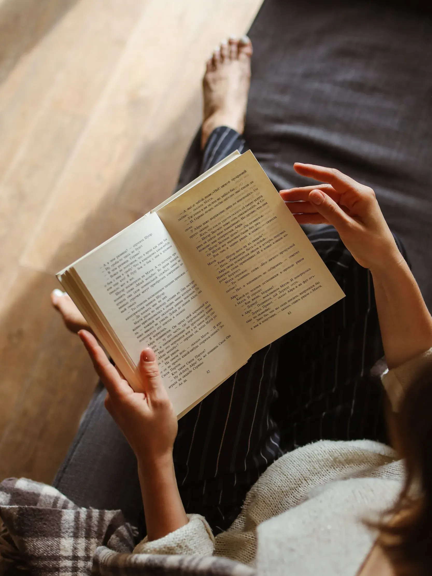 Una mujer descalza lee un libro tumbada en el sofá, con una manta de cuadros sobre las piernas