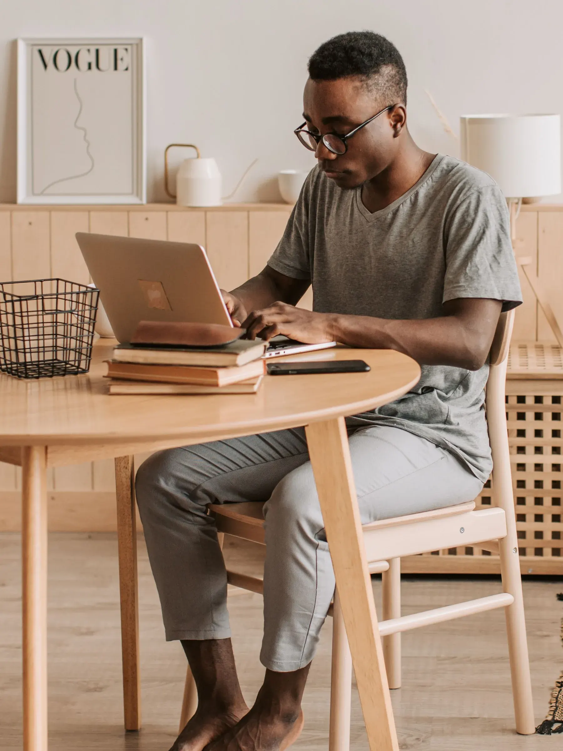 Un hombre con gafas escribe en su portátil sentado a una mesa de madera, con el móvil y unos libros al lado