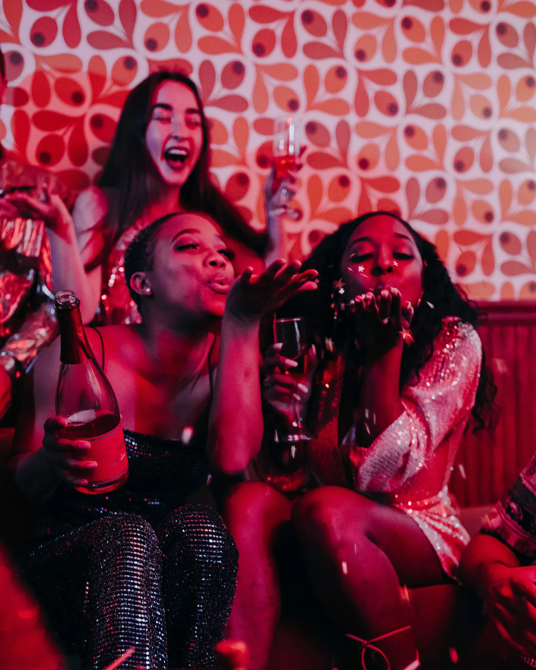 Tres amigas celebran con una botella y copas en un sofá, lanzando besos a la cámara